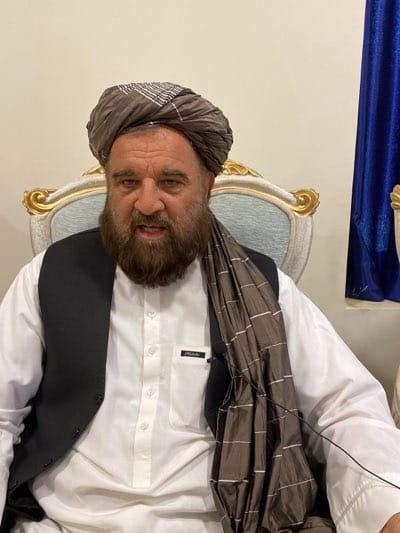 A man with a full beard and a distinctive turban is seated in an ornate chair. He wears a white shirt, a black vest, and a brown scarf. A small microphone is clipped to his shirt. Behind him is a blue curtain, partially visible. The scene suggests an indoor setting, possibly a room in Arghandab, Afghanistan, given the prompt. No specific actions or interactions are apparent.