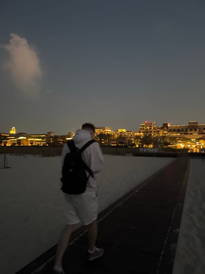 A male individual is depicted walking away from the camera along a dark wooden boardwalk on a sandy beach at dusk. He wears a white hooded sweatshirt, white shorts, and light-colored sneakers, and carries a black backpack. His hair is short and light-colored. The boardwalk extends into the middle distance, flanked by light-colored sand. In the background, a series of multi-story buildings, illuminated with warm yellow lights, line the horizon. These buildings feature distinctive architectural elements consistent with luxury resorts. The sky above is a dark blue-grey, with a single prominent white cloud in the upper left quadrant and a small, faint white point of light in the upper right. The scene is located on Saadiyat Island, Abu Dhabi, United Arab Emirates, consistent with the architecture of beachfront resort properties in that area.