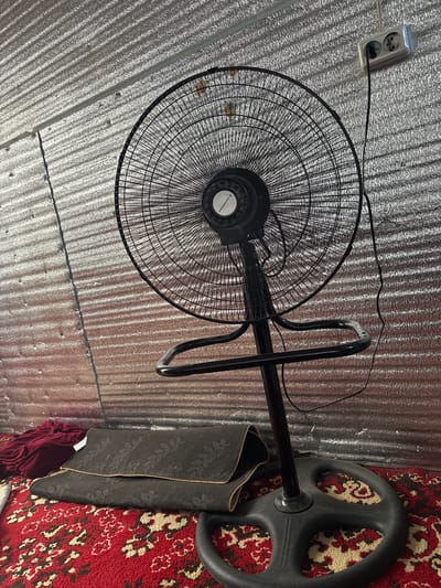 A black oscillating fan stands in a room with a metallic, corrugated wall and a red, patterned carpet. A dark-colored mat is folded in front of the fan. To the left of the mat, a burgundy piece of fabric is visible. A power cord from the fan is plugged into a double electrical outlet mounted on the wall.