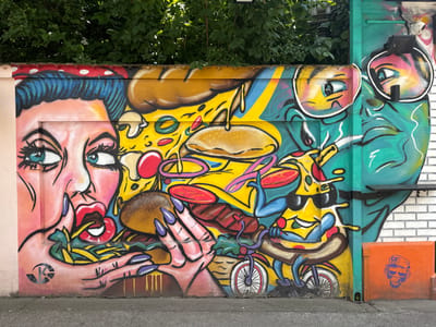 This image depicts a vibrant mural on a wall in Sremska Mitrovica, Serbia. The artwork features a stylized representation of food and figures in a pop-art style. On the left, a woman's face with blue hair, large blue eyes, and red lips is depicted, seemingly interacting with a large hamburger. To the right, a segment of a green face with large sunglasses is visible, and further to the right, a pizza slice character wearing sunglasses rides a miniature bicycle, holding a hot dog. The mural is rich in color, with prominent yellows, reds, blues, and greens. The wall itself appears to be constructed of concrete blocks, with some brickwork visible on the far right. Overhanging the wall is a dense canopy of green leaves from a tree. The ground in front of the mural is asphalt. An orange box with a stencil of a monkey head wearing a cap is positioned at the base of the wall on the right. The mural has a playful and energetic feel, likely intended as a visual attraction in the urban landscape of Sremska Mitrovica.