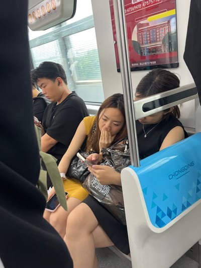 The image depicts passengers on what appears to be a public transit vehicle, possibly a subway or train, with a sign indicating "CHONGQING" visible on a blue and white panel. The scene is indoors, with large windows showing a blurred outdoor environment. In the foreground, three individuals are seated. A man on the left, wearing a black t-shirt and earbuds, is looking down at his phone. To his right, a woman in a yellow top is holding a phone and covering her mouth with her hand. Next to her, another woman in a black top is also looking down at her phone, wearing a necklace. There are bags and personal items visible, including a black quilted handbag and a clear plastic bag containing items. A sign with Chinese characters and an image of a building is visible on the upper right side of the image, along with a phone number. The overall impression is a typical scene of people commuting, engaged with their mobile devices.
