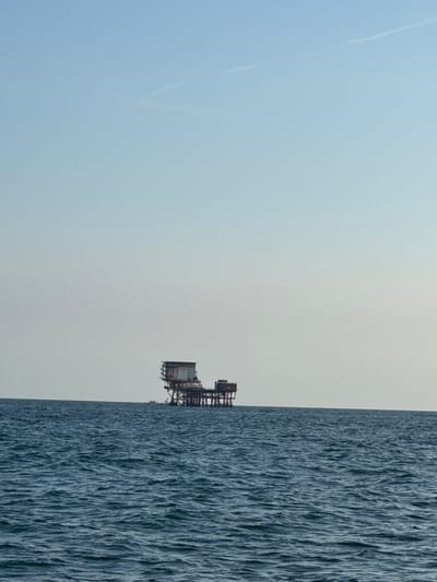 The image depicts an offshore platform situated in the open sea under a clear, pale blue sky. The platform is a multi-level structure with visible scaffolding and buildings on top. The sea is a deep blue with moderate waves. There is no indication of the city of Ravenna, Italy, in the image.