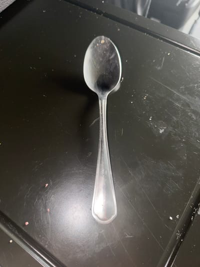 The image shows a single, metallic spoon lying horizontally across a dark, textured surface. The spoon has a polished finish and appears to be a standard eating utensil, possibly a teaspoon or dessert spoon. Its bowl is slightly curved and reflects the surrounding light, indicating it is clean or at least recently wiped. The handle of the spoon has a subtle, decorative rim at its end.

The surface beneath the spoon is a matte black, with a noticeable pattern of fine scratches and scuffs, suggesting it might be a countertop, a tray, or a similar utilitarian surface that has seen regular use. There are also a few small, light-colored specks scattered across the black surface, as well as some faint pinkish smudges. These details suggest the environment is not perfectly sterile and may be a kitchen or dining area where food preparation or consumption has occurred.

The lighting in the image is somewhat diffused, casting a gentle sheen on the spoon and the surface. There are no people visible, nor any other objects that would provide further context about the specific activity or event. The time of day and weather are not discernible from the image. The overall impression is a simple, straightforward depiction of a common household item in its environment, with no strong emotional cues or specific narrative implied. The location context of Union, United States of America, does not offer any specific visual cues within the image itself.