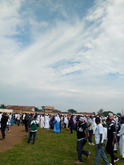 A large gathering of people are assembled in a field in Beni, Democratic Republic of Congo, located at coordinates 0.489 latitude and 29.463 longitude. The field is covered in short green grass. The sky is mostly cloudy with some areas of blue sky visible. The people are dressed in a variety of clothing, including traditional African garb and western attire.  There are many people wearing white robes and headscarves. Many people are looking around and interacting with each other. There are some buildings visible in the background, including a one-story building with a yellow sign.  Some people are walking away from the camera, and some are walking towards the camera.  There is a man wearing a green jacket and black pants walking to the left of the image, and a man wearing a black jacket and black pants walking to the right of the image.  There are many trees in the background. There is a small group of people gathered in the lower right corner of the image.  The atmosphere appears peaceful and celebratory.