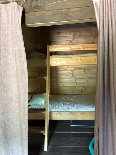 The image shows a close-up of a bunk bed in a wooden room. The bunk bed is made of light-colored wood and has mattresses on both the upper and lower bunks. There are light brown curtains on the left and right sides of the frame, partially obscuring the view of the room. The walls and ceiling are constructed of dark wooden planks. A green coiled hose is visible on the floor to the right, partially hidden by the curtain. The scene appears to be in a rustic setting, possibly a cabin or a dorm room. The location is identified as Bol'shaya Rechka, Russia. There are no people or animals visible in the image.