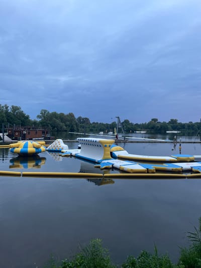 The image shows a water sports facility on a cloudy day, likely in the morning or late afternoon. The scene is set on a calm body of water, possibly a lake or a wide river. In the foreground, a large, inflatable water obstacle course is partially submerged, with yellow and blue sections. These inflatable structures are designed for recreation and adventure.
Behind the inflatable obstacles, several long, narrow boats, likely rowing shells, are docked or in the water. A few people are visible near the boats and on a wooden structure that appears to be a dock or a boathouse. One of the buildings on the dock has text that reads "WAKE PORT HOUSE BOAT". In the background, a densely wooded area lines the far bank of the water, suggesting a natural or park-like setting. A crane or similar structure is visible near the dock. The sky is overcast, with a soft, diffused light, indicating a lack of direct sunlight. The water's surface is smooth, reflecting the sky and the structures on the water. The overall impression is of a tranquil, slightly overcast day at a water recreation area.