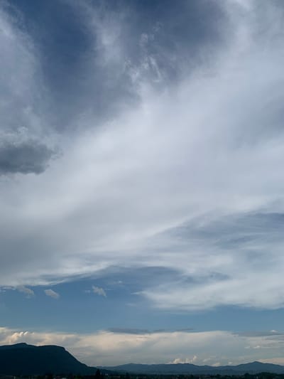 This media file depicts a wide-angle view of a sky with varied cloud formations. On the left side of the image, darker, more textured clouds suggest an impending change in weather or a more turbulent atmospheric condition. These clouds transition towards the right into lighter, wispy formations that stretch across the frame. The right side of the image reveals a strip of land, likely a distant mountain range or hills, silhouetted against the sky. The lighting suggests it is either early morning or late afternoon, with the sun casting a soft glow that illuminates the edges of some clouds. The overall scene is one of natural beauty, focusing on the dynamic interplay of light and cloud. There are no people, objects, or visible text in the frame. The location is identified as Khust, Ukraine.