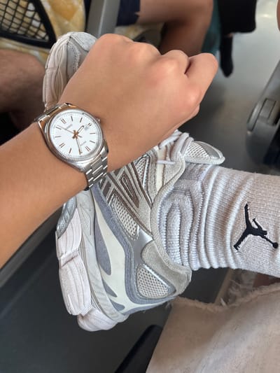 The image shows a close-up of a person's left hand wearing a silver watch with a white face and rose gold hands and hour markers. The watch band is also silver. The hand is resting on a light grey athletic shoe with white accents. The shoe is a New Balance 990v5, judging by the distinctive silhouette and sole design. The person is wearing light grey socks with a black Jumpman logo, associated with the Jordan brand, on the calf. The background is blurred but appears to be indoors, possibly on public transport given the metallic element at the top left and the suggestion of seating. The lighting suggests daytime. The overall mood is casual. No visible text other than on the watch face, which reads "CASIO" and what appears to be "5334" and "WR" (likely indicating water resistance). There is also a date window showing "12".