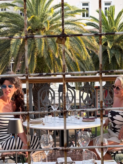 This image captures an outdoor dining scene, likely in Split, Croatia, viewed through a rustic metal grate. Two women are seated at a round marble-topped table with wicker chairs. The woman on the left has long, dark, curly hair and wears sunglasses and a striped dress. The woman on the right has blonde hair and sunglasses, and is wearing a black and white patterned top and a light-colored skirt. Palm trees are visible in the background, suggesting a warm, Mediterranean climate. The table is set with glasses, napkins, and what appear to be plates. A small lamp sits on the table to the left. The scene is bathed in warm, golden sunlight, creating long shadows. The presence of palm trees and the outdoor dining setup strongly suggest a location in a coastal city like Split.