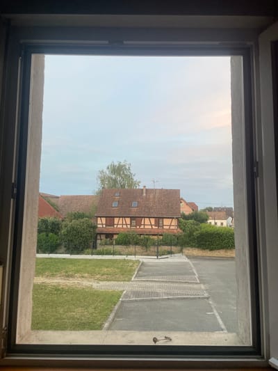 This image, taken from a window, shows a view of buildings and sky in Gerstheim, France. In the foreground, the window frame is visible, with a latch on the bottom sill. Beyond the window, there is a paved area and a grassy lawn. Further in the background, several houses with terracotta roofs are visible, nestled amongst trees. The sky is pale blue with some wispy clouds. The overall impression is a quiet, residential scene.