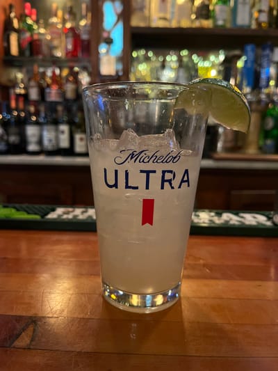 A Michelob Ultra-branded glass, filled with a light-colored beverage and ice, is garnished with a lime wedge on the rim. It sits on a wooden surface, likely a bar top, with a green and white patterned edge visible nearby. In the blurred background, a variety of bottles are displayed on shelves, suggesting a bar or restaurant setting. The lighting is soft and creates a warm, inviting atmosphere.