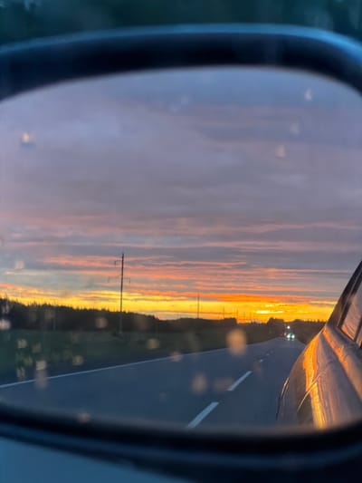 The image is a photograph taken from the perspective of a vehicle's side-view mirror, reflecting a sunset scene. The mirror itself, with its dark frame, occupies the left portion of the image, while the reflection fills the majority of the frame.
The reflection shows a road stretching into the distance. On the left side of the road, there is a vibrant sunset with hues of orange, yellow, and pink stretching across the sky. Wispy clouds are visible, catching the colors of the setting sun. Several utility poles with wires are spaced along the side of the road, partially silhouetted against the bright sky. To the right of the road, a dark landscape is visible, with hints of greenery and possibly trees. The road itself is asphalt and has white lane markings. A few distant vehicle lights can be seen on the road, suggesting traffic. The overall impression is that of a scenic drive during sunset. The time of day is clearly sunset. The location cues, such as utility poles and the type of road, suggest a rural or highway setting. The colors of the sunset evoke a feeling of peace and beauty. There is no visible text in the image.