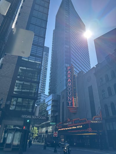 The image captures a vibrant, sunlit urban street scene, likely taken during the day, looking upwards towards tall buildings. The bright sun is positioned near the top of the frame, creating a prominent lens flare and illuminating a modern glass skyscraper that reflects the clear blue sky.
In the mid-ground, a classic theater marquee for the "PARAMOUNT" is prominently displayed, featuring its name in large, illuminated red letters against a dark background, accented with numerous yellow lights. Below this, a digital marquee advertises a performance: "Mac and Matt Ray. Songs from Bark of Millions :: OCT 15-18". Further up, parts of other building signs are visible, including "NORMANDIE BALLROOM" and "BIJOU".
At street level, a large vertical sign for "LEGAL SEA FOODS" is visible on the side of a building. Street infrastructure, such as lampposts, traffic lights, and street signs, are also present. One street sign clearly indicates "AVENUE DE LAFAYETTE", and a "DO NOT ENTER" sign is also visible. The overall impression is a bustling city environment on a clear, bright day, with no discernible people but clear signs of urban activity. The combination of the Paramount Theatre, Legal Sea Foods, and Avenue de Lafayette strongly indicates the location is Boston, Massachusetts, United States.