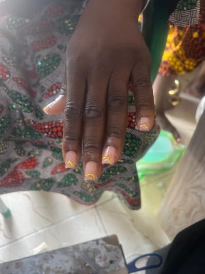 A close-up, high-angle shot shows a person's left hand with light pink nails adorned with a gold French tip design. The hand is positioned in front of a patterned fabric, likely clothing, featuring red, green, and white circular motifs. To the right of the hand, a gold hoop earring is visible on another person's arm, and further in the background, a bright yellow and orange patterned fabric is partially visible. A light green plastic container is also present in the mid-ground. The scene appears to be indoors, possibly in Wauro Jabbe, Nigeria, given the context of the provided information.