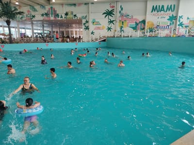 The image shows a busy indoor aquapark with a large wave pool. Numerous people, including adults and children, are swimming and playing in the bright turquoise water. Some individuals are using inflatable rings or vests. The aquapark features a decorative mural on the wall depicting a cityscape with palm trees and tropical motifs, along with the text "MIAMI AQUAPARK & SPA" and "ЗАРЯДНАЯ СТАНЦИЯ" (Charging Station). The environment appears to be a recreational facility. This aquapark is located in Irgelі Rural District, Kazakhstan.