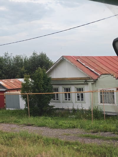 A weathered, light green, wooden house with a bright red, corrugated metal roof stands in a rural setting. The house has white trim around its windows, which are multi-paned. A dark wire runs diagonally across the overcast sky above the house. In front of the house, a rusty metal fence with orange poles encloses a grassy yard. To the left of the main house, a smaller structure with a similarly red, corrugated metal roof is visible. The ground in front of the property is a mixture of gravel and grass. The scene is likely located in the village of Kuvak-Nikolskoye, Russia, given the architectural style and surrounding vegetation.