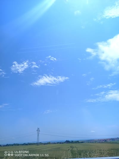 The image displays an expansive outdoor landscape in Krustina, Bulgaria, captured on a bright, clear day. The upper three-quarters of the frame are dominated by a vibrant blue sky with scattered white cumulus clouds; a strong lens flare or sunbeam originates from the upper-left quadrant.

The horizon line bisects the lower quarter of the image, separating the sky from the terrestrial environment. The mid-ground features vast, undulating green agricultural fields. A single metallic electrical transmission tower (pylon) stands prominently in the mid-left, supporting multiple power lines that traverse the scene horizontally. In the distant mid-right, a village settlement is visible, characterized by numerous buildings with reddish-brown roofs nestled amongst greenery.

The immediate foreground reveals dense, low-lying green and dry vegetation, with a partial metallic guardrail or barrier visible along the very bottom edge, particularly towards the center and right. There are no individuals, animals, or specific actions depicted in the scene. A digital watermark, "ULEFONE SHOT ON ARMOR 11 5G," is present in the lower-left corner.