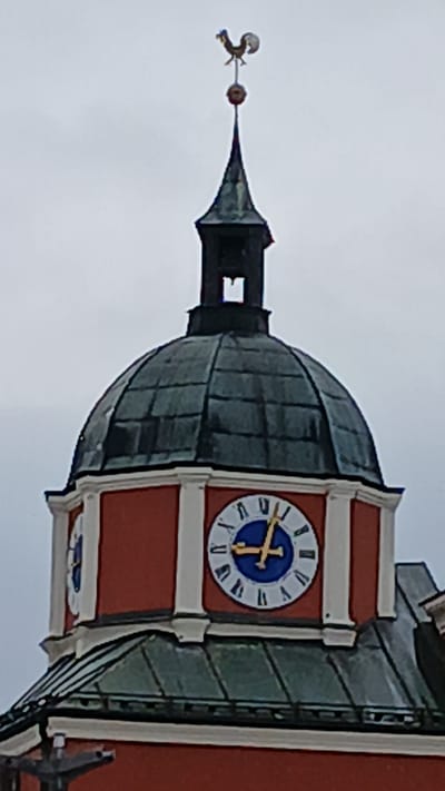 The image shows the upper section of the bell tower of the St. Jakobus church in Pfarrkirchen, Germany. The tower is topped with a weather vane shaped like a rooster. Below the weather vane is a dark spire, and then a red octagonal section featuring two clock faces. The clock faces have Roman numerals and blue and gold hands. The lower part of the image shows a green tiled roof section. The sky is overcast.
