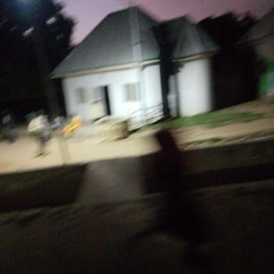 The image captures a blurry outdoor scene at dusk in Wuro Allahamdu, Nigeria. In the background, a light-colored building with a dark roof is partially visible. The building appears to have windows and possibly a ramp or walkway leading to its entrance. To the left of the building, dark trees are present. The foreground is dominated by a dark, out-of-focus area, possibly a pathway or ground surface. In the mid-ground, there are blurred shapes that suggest human figures and possibly playground equipment, indicated by a yellow object. The overall image quality is poor due to blurriness and low light conditions, making precise identification of all elements difficult.