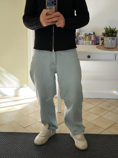A person stands indoors, holding a phone, and wearing a black zip-up sweater, light blue jeans, and white sneakers. The person is positioned between a white wall with a shelf and decorative items (plant, books, wicker basket) and a staircase leading to a floor with beige diamond-patterned tiles and a dark gray rug. Light is entering the room from the right side. The location is not evident in the provided image.