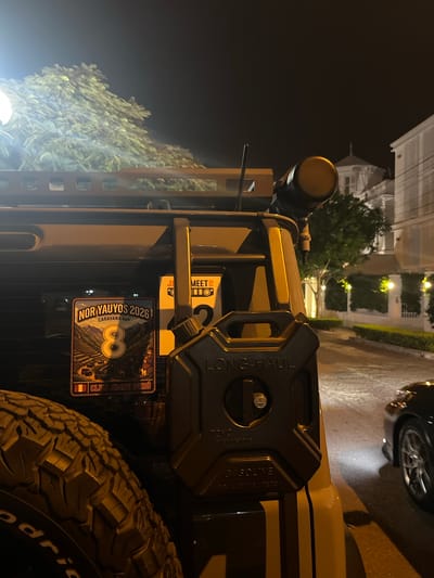 A nighttime shot reveals the rear of a vehicle parked in the Barranco district of Lima, Peru. The "NOR YAUYOS 2026" sticker, representing a 4x4 caravan, is prominent, alongside a sticker from "CLUB JIMNY PERÚ", indicating the car's affiliation and origin. A black "LONG-HAUL" gasoline container is mounted on the vehicle's rear. A partially visible tire with "odric" visible in a white font occupies the right side. An additional sticker is placed nearby on the vehicle. In the background, streetlights illuminate the wet road and colonial architecture of Barranco, including the tops of buildings, trees, and parked cars.