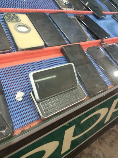 The image displays a collection of smartphones and mobile phones laid out on a blue, perforated mat with an orange border, likely in a market or shop. A variety of modern smartphones with large screens are visible, mostly black. In the foreground, a classic Nokia phone with a slide-out QWERTY keyboard is prominently featured. The Nokia phone's screen displays the word "NOKIA" and a model number "E7". The keyboard has alphanumeric keys and symbols.
To the left, a yellow-cased smartphone with a prominent camera module and a circular cutout is visible. Text on this phone appears to read "IP 13 Pro". Several other black smartphones are arranged behind and to the right of the Nokia. Some of these appear to be disassembled or have damaged screens. The setting suggests a retail environment where mobile phones, both new and possibly used or for repair, are being sold or displayed. The lighting is bright, indicating it's likely daytime. The background shows a green sign with stylized cream-colored text, partially obscured. The overall scene is of a display of mobile technology.