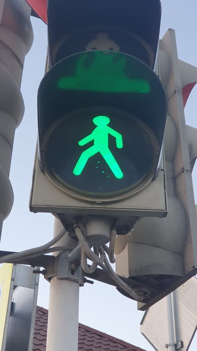The image shows a close-up of a pedestrian traffic signal displaying a green walking person symbol. The signal is mounted on a metal pole with various wires and conduits attached. Above the green pedestrian signal, a traffic signal housing is visible, and a red triangular sign is partially seen at the top left. In the background, a clear sky and a tiled roof of a building are present, suggesting an outdoor urban environment. The image does not contain any people or specific landmarks that would identify the exact location within Ust-Labinsk, Russia. The lighting suggests it is daytime, possibly late afternoon or early morning due to the angle of the light.