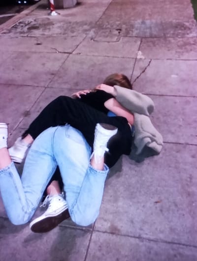In Mirbozor, Uzbekistan, two individuals are observed lying on a paved sidewalk under artificial light. One person wears light blue jeans and white low-top sneakers with brown soles. Their body is partially obscured by the second individual, who is positioned on top and embracing them. This second individual wears a black long-sleeved garment and a light grey hooded sweatshirt, with the hood partially covering their head. Their arms are wrapped around the first person. The ground is a light grey paved surface with visible seams and cracks. In the blurred background, elements consistent with an urban environment, including a partially visible red and white striped bollard-like object, are discernible.