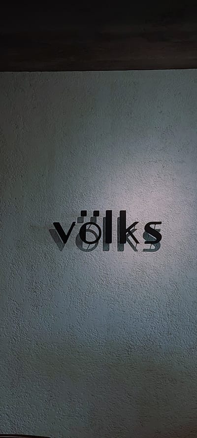 The image displays the word "völks" in a stylized, dark font, appearing to be a sign or logo, mounted on a textured, light-colored wall. The letters cast shadows on the wall, indicating a light source from above and to the left. The upper portion of the image shows a dark, horizontal element, possibly a ceiling or a shelf, casting a shadow on the wall below it. The wall has a rough, granular texture. The scene is devoid of people and appears to be an indoor or covered outdoor setting. The location in Cicalengka, Indonesia is not discernible from the visual elements within the image itself.