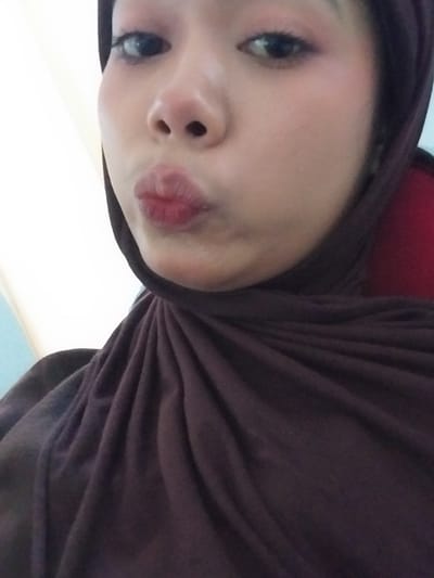 The media file is a close-up, low-angle, portrait photograph of a young person wearing a dark brown hijab. The subject is looking towards the camera with their lips puckered, as if blowing a kiss. Their right eye, visible on the left side of the frame, is open and appears to have eyeliner and mascara applied. The left side of their face is mostly obscured by the hijab, but their cheek and nose are visible.

The subject is wearing a dark brown hijab, which drapes around their head and shoulders. A hint of a red fabric is visible underneath the hijab on the top left of the image. The background is out of focus and appears to be an indoor setting, with a light-colored wall or surface dominating the lower portion of the frame and a faint blue on the far right.

There are no visible text elements in the image. The lighting suggests it was taken indoors, possibly during the daytime given the brightness of the background. The subject's expression is playful and affectionate. The image is oriented such that the top of the head is towards the left.