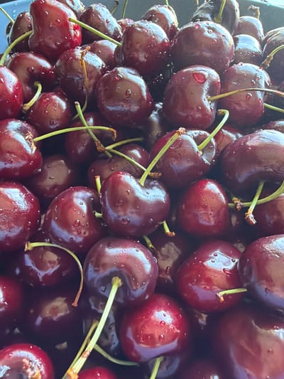 The image displays a close-up, overhead view of a large pile of dark red cherries, many of which are glistening with small water droplets. The cherries are ripe and appear plump, with some still attached to their stems. The lighting suggests it might be daytime, with natural light illuminating the fruit. The texture of the cherries is smooth and glossy, and the vibrant red color is prominent throughout the frame. The overall impression is one of freshness and abundance, possibly indicating a harvest or a market stall. There is no visible text in the image.