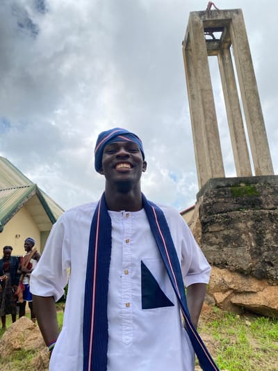 The image depicts a young Black man smiling at the camera, wearing a white tunic and a blue and white striped headwrap, in front of a stone monument. He has a blue and white striped sash draped around his neck. The monument is tall and appears to be made of concrete, with open sections near the top. In the background, to the left of the man, is a building with a green, sloped roof. To the far left, two other individuals are visible, dressed in traditional attire. The ground is covered with grass and scattered rocks. The sky is cloudy and overcast. The location is identified as Wang, Nigeria.