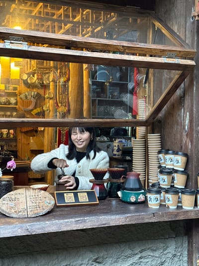 The scene depicts a woman engaged in beverage preparation at an open-air service window within a building in the city of Gongtan, China. The window is constructed of weathered wood, with its two glass panes hinged at the top and angled upwards and outwards. An upper glass section of the window reveals interior wooden beam construction. A smiling woman with dark hair and bangs, wearing a cream-colored textured cardigan over a dark top, operates a manual coffee grinder with both hands from behind the counter.

On the wooden counter, from left to right, are: an oval wooden menu board with inscribed Chinese characters; a glass jar containing dark contents, likely coffee beans; a small, reddish-brown bowl; and a single purple orchid bloom. Central to the counter are two square QR code displays mounted on a wooden stand. To their right, two pour-over coffee drippers (one red/black, one dark green/brown) are positioned alongside a dark ceramic kettle resting on a teal-colored warming base. Further right, a tall stack of plain white paper cups stands beside multiple stacks of smaller, lidded kraft paper cups, several of which bear the Chinese characters '龚滩'.

The interior visible behind the woman features rustic wooden paneling and structural posts. Shelves hold an assortment of decorative and utilitarian objects, including various ceramic mugs and bowls, traditional folding fans (some with characters), and colorful strung ornaments. The exterior wall beneath the counter is constructed of rough-hewn stone blocks.