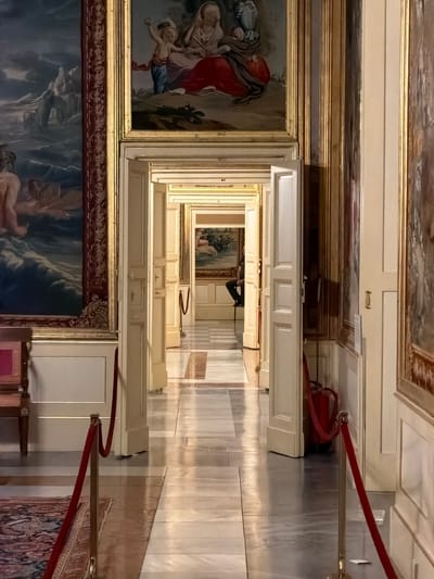 The image depicts a long, ornate hallway within a grand building, likely a palace or museum. The hallway is lined with large, elaborately framed paintings displayed on the walls. The architecture features classic design elements, including tall, white-paneled doors and high ceilings with decorative moldings. Sunlight streams in from unseen windows, casting long shadows and highlighting the polished marble floors, creating a dramatic and somewhat disorienting perspective due to the camera's vertical orientation.

In the foreground, a large, dark-framed painting is partially visible, featuring what appears to be a mythological or biblical scene with dramatic lighting and figures. To the left, another framed artwork shows figures in rich red and blue robes, possibly a religious depiction. Further down the hallway, more paintings are visible, receding into the distance, creating a sense of depth and grandeur.

A red velvet rope barrier, supported by brass stanchions, runs along the right side of the hallway, guiding visitors and indicating that the area may be an exhibition space. In the mid-ground, a fire extinguisher is mounted on the wall near a doorway, a practical safety detail within the opulent setting. The overall impression is one of historical significance and artistic wealth, likely within a well-preserved cultural heritage site. The lighting suggests it is daytime, and there are no indications of weather. The scene evokes a sense of quiet contemplation and admiration for the art and architecture.