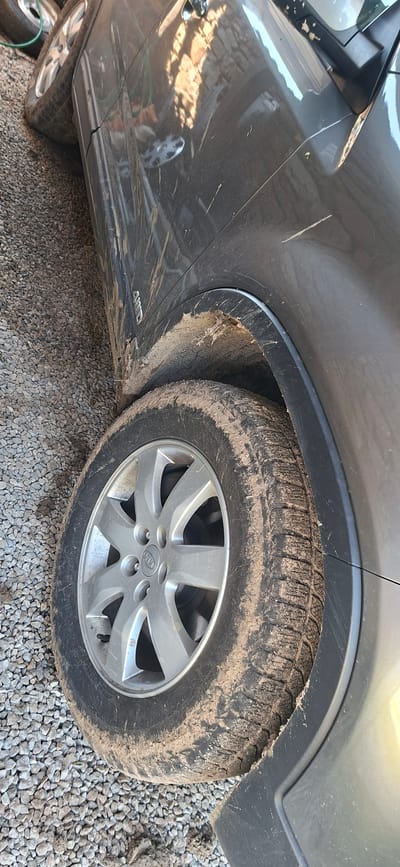 The image displays the side profile of a dirty, dark gray Kia SUV, parked outdoors on a gravel surface. The vehicle is heavily soiled with mud and dust, particularly on the front passenger-side tire, its alloy wheel, and the surrounding wheel well and lower body panels. The tire exhibits a distinct, chunky tread pattern, typical of all-season or winter tires, coated in dried mud. The silver alloy wheel prominently features the "KIA" logo on its hubcap.

Evidence of recent travel through muddy terrain is clear from the significant dirt splatters and streaks across the car's paintwork, extending from the tire up the fender and door panel. Several visible scratches, including a distinct white line on the fender and smaller marks near the door handle area, suggest wear and tear. Reflections on the car's glossy surface reveal glimpses of a nearby structure, possibly a building with brickwork, and bright daylight conditions.

The ground beneath the car is composed of small, light gray gravel stones, interspersed with patches of accumulated mud and dirt. A partial view of another tire or wheel is visible in the bottom left corner, suggesting another vehicle or a spare wheel. The "AWD" badge is clearly visible on the lower part of the car's side panel, indicating its all-wheel-drive capability. The scene captures the vehicle in a state reflecting rugged use or recent off-road driving.