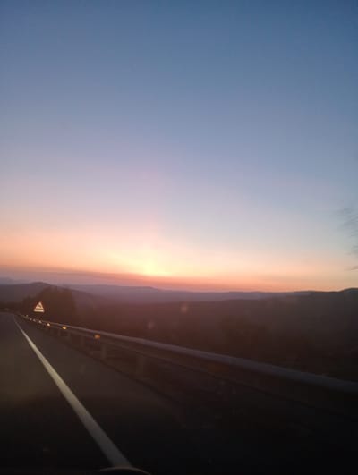 The image depicts an outdoor scene near Casas del Puerto, Spain, during twilight or dawn. The sky presents a gradient from a deep blue at the zenith to a vibrant orange and pink along the horizon. Silhouetted against the bright horizon are multiple layers of dark, undulating mountain ranges. In the foreground, a dark asphalt road extends into the distance, marked by a solid white line on its left side. A metallic guardrail, fitted with yellow/orange reflectors, parallels the road on the right. A white triangular warning sign with a red border, indicating a winding road and the text "1000 MT," is visible on the left shoulder, partially obscured by dark foliage. The scene is devoid of individuals. The perspective suggests the image was captured from inside a moving vehicle.