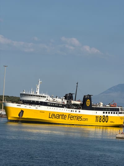 A yellow and white ferry, the "Kefalonia," is docked in a harbor. The ferry is large and appears to be a car ferry, with multiple decks visible. The word "Kefalonia" is written in black on the yellow hull, along with "Levante Ferries.com" and the number "11880". A website address, "vrisko.gr", is also visible. In the background, there are trees and hills. The sky is blue with some clouds. A single person is visible on a small dock to the right of the ferry, appearing to be fishing. The water is a deep blue. The scene is set in the Municipal Unit of Kastro - Kyllini, Greece.