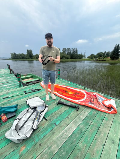 A man stands on a weathered green wooden dock by a lake in Nikol'skoe, Russia. He wears a green t-shirt, denim shorts, a black baseball cap, and white sneakers. He holds a black flexible hose in his hands. In front of him on the dock are several items: a deflated red and orange stand-up paddleboard with a dragon design, a black air pump, a large grey bag containing badminton rackets and a paddle, a green duffel bag, and two fishing rods. A small green rowboat is partially submerged near the dock. The lake is calm with reeds growing along the bank, and trees line the distant shore under a cloudy sky.