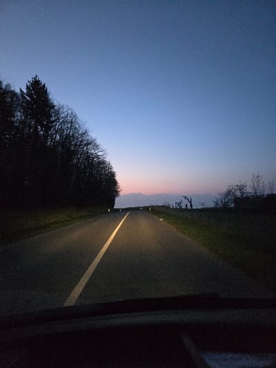 The image captures a rural road scene at twilight, originating from Hautemorges, Switzerland. The perspective is from within a vehicle, with the lower segment of the frame showing the vehicle's dashboard or windshield base. The sky above transitions from a deep blue at the zenith to a lighter, subtle pink-orange glow near the horizon, where a distant, silhouetted mountain range is visible. The asphalt road, dark gray in color, extends straight into the distance, illuminated directly ahead by two distinct beams from the vehicle's headlights. A continuous white line marks the center of the road. To the left, a dense mass of dark, silhouetted trees, including both coniferous and deciduous species, forms a forest edge. To the right, a dark grassy field extends, featuring a few sparse, bare-branched trees and some indistinct, distant structures. Small, reflective road markers are visible along both edges of the road, catching the headlight illumination. No individuals or specific actions beyond implied vehicular movement are discernible.