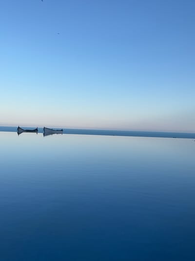 The image depicts an infinity pool with a calm, reflective surface that merges seamlessly with the horizon. Two small boats, rendered in a light color and appearing somewhat aged, are positioned on the water's edge. The sky above is a clear, gradient blue, transitioning from a lighter hue near the horizon to a deeper shade at the top. A few distant birds are visible in the sky. The overall scene conveys a sense of tranquility and vastness, with the reflective water creating a mirror-like effect that doubles the visual of the boats and the sky. The location is identified as Topola, Bulgaria.