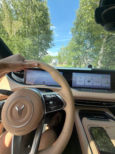 A first-person perspective shot captures a hand on the steering wheel of a car. The interior of the car is visible, featuring a large digital display dashboard and a tan leather steering wheel with an emblem. The car is on a road lined with trees, and houses are visible in the distance. The sky is blue with scattered clouds. The car appears to be traveling through a rural area, possibly on a summer day, given the lush greenery. The scene suggests a typical driving experience in a suburban or rural setting in Malinovka, Russia.