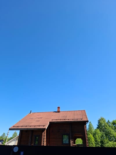 This image, taken in Kondratovo, Russia, captures a bright, sunny daytime scene dominated by a vast, clear blue sky. A robust wooden house with horizontal log walls and a reddish-brown tiled roof occupies the right side of the frame. A small chimney is visible on the roof. The house features windows, one reflecting green foliage, and a distinctive, possibly arched or circular opening on its facade.
Lush green trees are prominent in the upper right corner, bordering the top of the house. A dark fence or wall runs along the lower right edge of the image, partially obscuring another light-colored building and utility poles with wires behind it. On this dark fence, a small blue rectangular sign with the number "60" is clearly visible. Below it, a white circular object is present, though no discernible text is on it. The weather is clear with strong sunlight, indicating a pleasant day. No people or specific activities are depicted; the scene is a tranquil architectural and natural landscape. A few faint, tiny dark specks, possibly birds or dust, can be observed against the expanse of the blue sky.