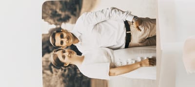 An outdoor photograph, possibly taken in the city of Winnenden, Germany, depicts a young couple posed together. The man, on the upper left, is wearing a white collared shirt, a black belt, and light-colored pants. He is looking towards the camera with a slight smile. The woman, on the lower right, is wearing a white dress and also smiling at the camera. Her arm is visible resting on a white fabric. The background appears to be a natural outdoor setting with trees and foliage. The image has a sepia-toned filter, giving it a vintage or artistic quality.