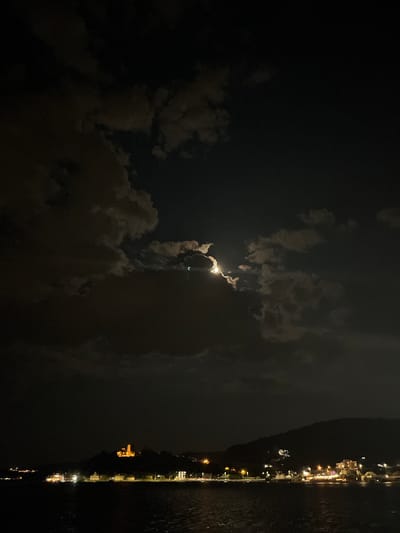 This is a vertical, nighttime photograph taken from a high vantage point, overlooking a body of water and a coastal town. The sky is dark with scattered clouds, some of which are illuminated by a bright, partially obscured moon. The moon appears to be low in the sky, and its light creates a glow around the edges of the clouds.
On the right side of the image, a town is visible along the coastline. Numerous lights from buildings and streetlights create a vibrant strip of illumination against the dark hillside. The lights reflect on the dark water, creating shimmering patterns. The town appears to be nestled on a steep slope, with lights climbing up the hillside. The overall impression is of a peaceful, illuminated coastal settlement at night.
The scene suggests a calm night, with the moon providing a dramatic backdrop to the illuminated town. The presence of clouds adds a dynamic element to the sky, partially hiding and revealing the moon. The image captures the beauty of a nocturnal landscape where natural light from the moon and artificial light from human settlement converge. There is no visible text in the image.