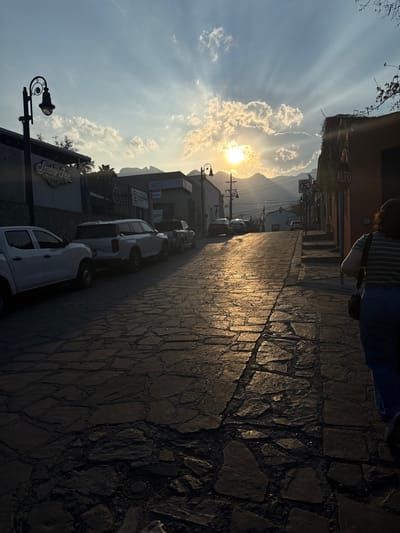 The image displays a sloped cobblestone street in Santiago, Mexico, captured during sunset. The low sun, positioned slightly right of center, radiates a bright golden light through scattered clouds, creating visible sunbeams and illuminating the upper half of the street. The foreground and the left side of the scene are cast in deep shadow.

On the left, a line of vehicles is parked uphill, including a white pickup truck and a white SUV closest to the viewer. A dark, ornate street lamp extends from the facade of a building immediately to the left. Further down the street on the left, a sign for "Smokey Joe's Mexican" is visible on a building, alongside other commercial or residential structures.

On the right side of the street, a person is walking uphill, viewed from behind. They are wearing a horizontal striped shirt, blue jeans, and a dark shoulder bag. Adjacent to this person is a building with an orange/terra cotta colored facade, featuring climbing vegetation. A red circular "ALTO" (STOP) sign is affixed to a pole near the right edge, with steps visible leading upwards along the building.

The street surface consists of irregular, dark-toned cobblestones, with the sun's reflection highlighting sections of the wet or polished stone. In the distant background, hazy mountains are visible, silhouetted against the bright sky. Utility poles and overhead wires are present in the mid-ground, completing the urban landscape.