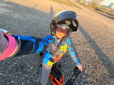 A person wearing a black motorcycle helmet, a blue and grey long-sleeved shirt with "Red Bull" and "FOX" logos, and grey leggings is lying on their back on a gravel surface. The person is positioned on or near an orange and black electric scooter or small motorbike. The individual appears to be holding a smartphone or camera to take a selfie. In the background, there is a glimpse of a cityscape, possibly indicating the location of Азьмушкино, Russia. The scene suggests an outdoor setting, possibly a roadside or recreational area.