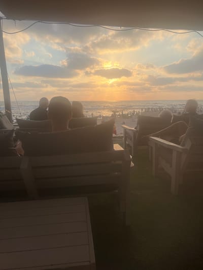 This media file captures a sunset scene at a beachside location, likely within Hof HaCarmel Regional Council, Israel. The foreground is dominated by the silhouettes of several people seated on what appear to be beach chairs, creating a sense of relaxed observation. They are positioned on a grassy area, with a white wooden structure, possibly a wall or partition, visible to the right.
Beyond the seated figures, the scene opens up to a beach lined with numerous beach chairs and umbrellas, indicating a populated public space. The ocean stretches out to the horizon, with gentle waves visible. The sky is filled with dramatic clouds, illuminated by the warm, golden hues of the setting sun. The sun itself is partially obscured by clouds, casting a radiant glow and creating shafts of light that pierce through the atmospheric haze.
The time of day is clearly late afternoon or early evening, as evidenced by the sunset lighting. The weather appears to be clear, with the clouds contributing to the scenic beauty rather than suggesting inclement conditions. The overall mood conveyed is one of tranquility and enjoyment of a beautiful natural spectacle. There is no visible text in the media file.