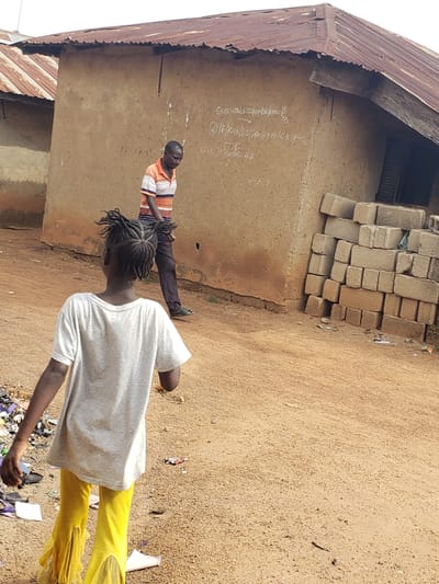 In Kachia, Nigeria, a dirt path leads towards a compound with buildings constructed of mud walls and corrugated metal roofs. In the foreground, a young girl with braided hair, wearing a grey t-shirt and bright yellow pants, walks away from the viewer. Behind her, a man in an orange and striped t-shirt and dark pants is visible, seemingly carrying or interacting with a bundle of dark, coiled material. To the right of the buildings, a stack of concrete blocks is piled against the wall. The ground is sandy and dry, with scattered debris. White writing is visible on the mud wall of the building.