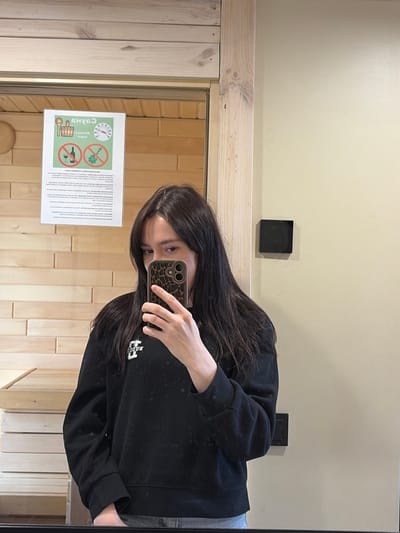 A person is seen taking a mirror selfie indoors. The subject is a female with long, dark hair, wearing a black hooded sweatshirt with a white logo (partially visible, possibly "BOJO"). She holds a smartphone with a distinctive leopard print case, obscuring her mouth and chin as she takes the photo, with her eyes looking towards the camera.
The setting appears to be a room, possibly within a spa or sauna facility, given the prominent light-colored wooden paneling on the wall, which has a distinct block pattern. Above the wooden paneling, the wall is painted a light, neutral beige color. A small, black rectangular object, possibly an electrical outlet or light switch, is mounted on the beige wall.
Reflected in the mirror, to the left of the person, is a white rectangular sign attached to the wooden wall. The sign features Cyrillic text at the top, including "САУНА" (SAUNA), confirming the location context as a sauna. Below the text are several pictograms: a thermometer, a bucket with a ladle, a crossed-out bath broom (venik), and a crossed-out bottle, likely indicating rules or recommendations for sauna use. The overall ambiance suggests an indoor, clean environment. The location is Мишкино, Russia, consistent with the Cyrillic text on the sign.