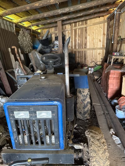The image depicts the interior of a rustic wooden shed in Бородулино, Russia. The shed is filled with various items, suggesting storage and possibly agricultural use. Dominating the foreground is a blue and black tractor, its front grill featuring two rectangular headlights and a fan visible behind a metal grille. The tractor is dirty, with mud caked onto its tires. To the left of the tractor, a collection of dried branches, possibly a bathhouse broom (venik), hangs from the ceiling. Various tools and implements, including a curved metal object resembling a handle, are leaning against the walls. The shed's construction is visible, with rough-hewn wooden planks forming the walls and ceiling, supported by wooden beams. The floor is cluttered with debris, including dirt, straw, and discarded materials. In the background, stacks of red bricks and buckets are visible, along with other indistinct objects. The overall impression is one of a functional, albeit somewhat disorganized, storage space in a rural setting. There are no people present in the image.